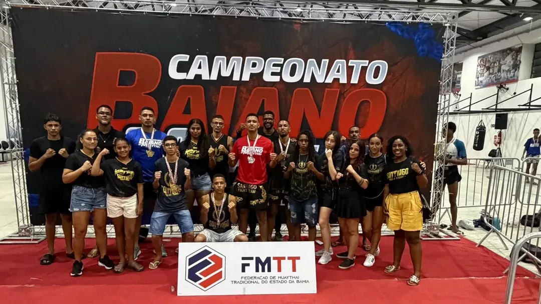 Equipe Santiago Fight Team reunida no Campeonato Baiano diante de painel oficial com placa da FMTT